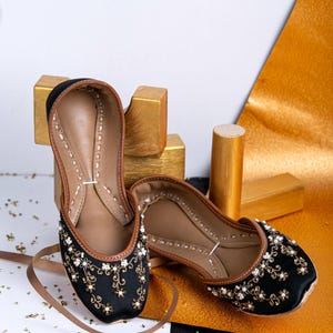 Pode incluir: Um par de sapatos baixos pretos bordados com detalhes dourados e brancos. Os sapatos est&atilde;o sobre uma superf&iacute;cie branca e preta com glitter dourado.