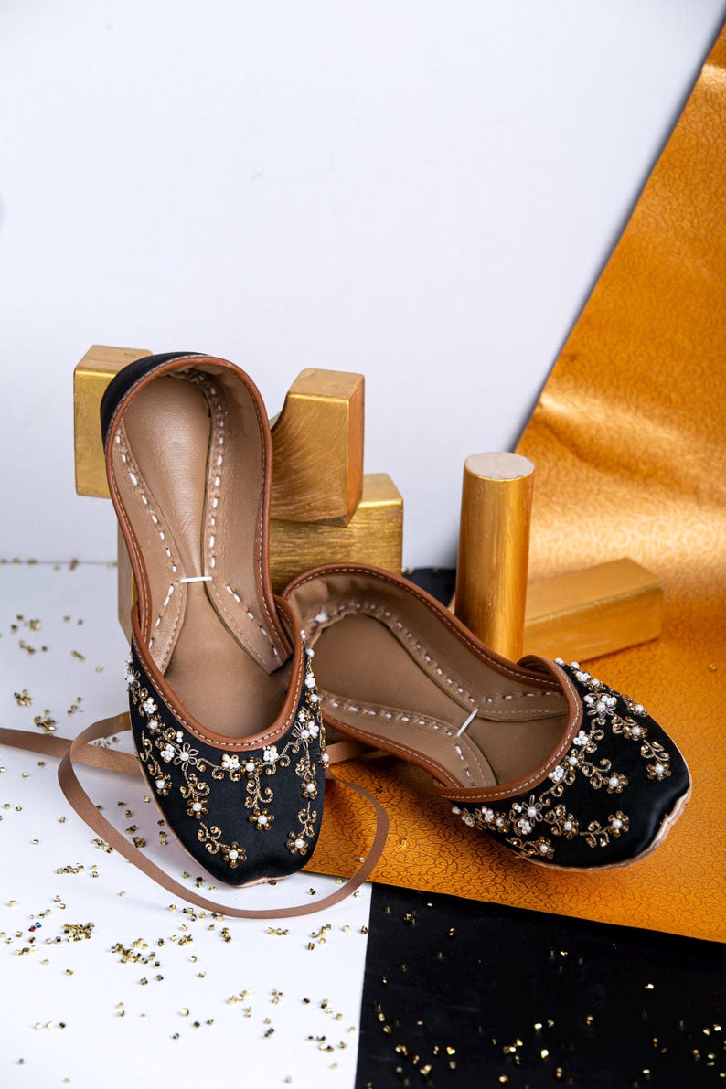 Pode incluir: Um par de sapatos baixos pretos bordados com detalhes dourados e brancos. Os sapatos est&atilde;o sobre uma superf&iacute;cie branca e preta com glitter dourado.