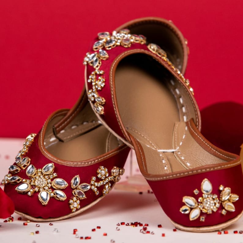 Puede incluir: Un par de zapatos de sat&eacute;n granate con intrincados adornos de oro y plata. Los zapatos tienen una punta puntiaguda y un tac&oacute;n bajo.