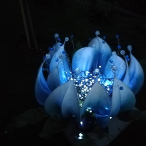 Pode incluir: Uma flor azul feita de tecido com uma estrutura de arame e luzes brancas dentro. A flor est&aacute; iluminada e brilhando.