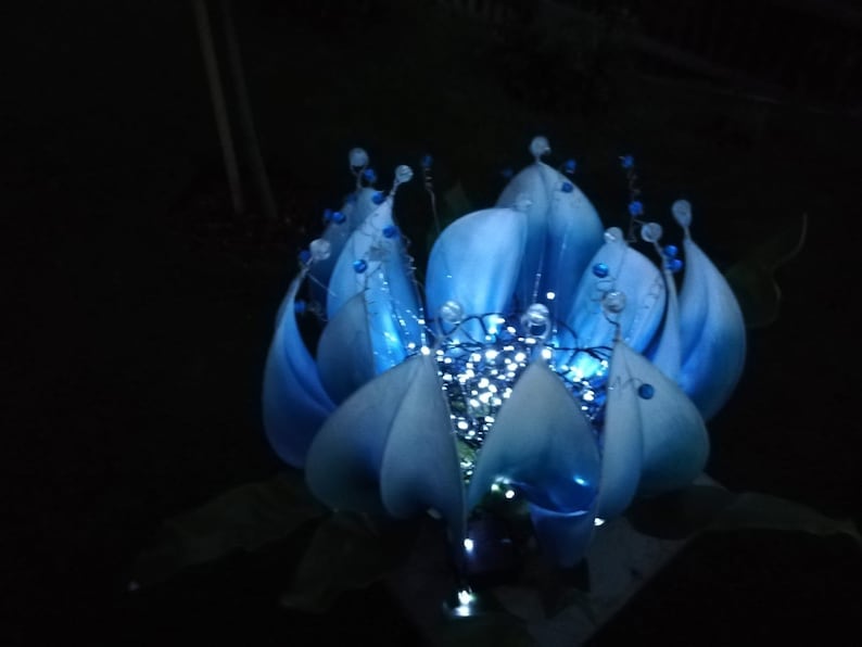 Pode incluir: Uma flor azul feita de tecido com uma estrutura de arame e luzes brancas dentro. A flor est&aacute; iluminada e brilhando.