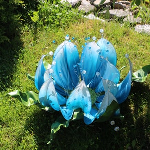 Pode incluir: Uma escultura de flor azul e branca com detalhes em contas. A escultura &eacute; feita de tecido e arame e est&aacute; sentada em um leito de grama verde.