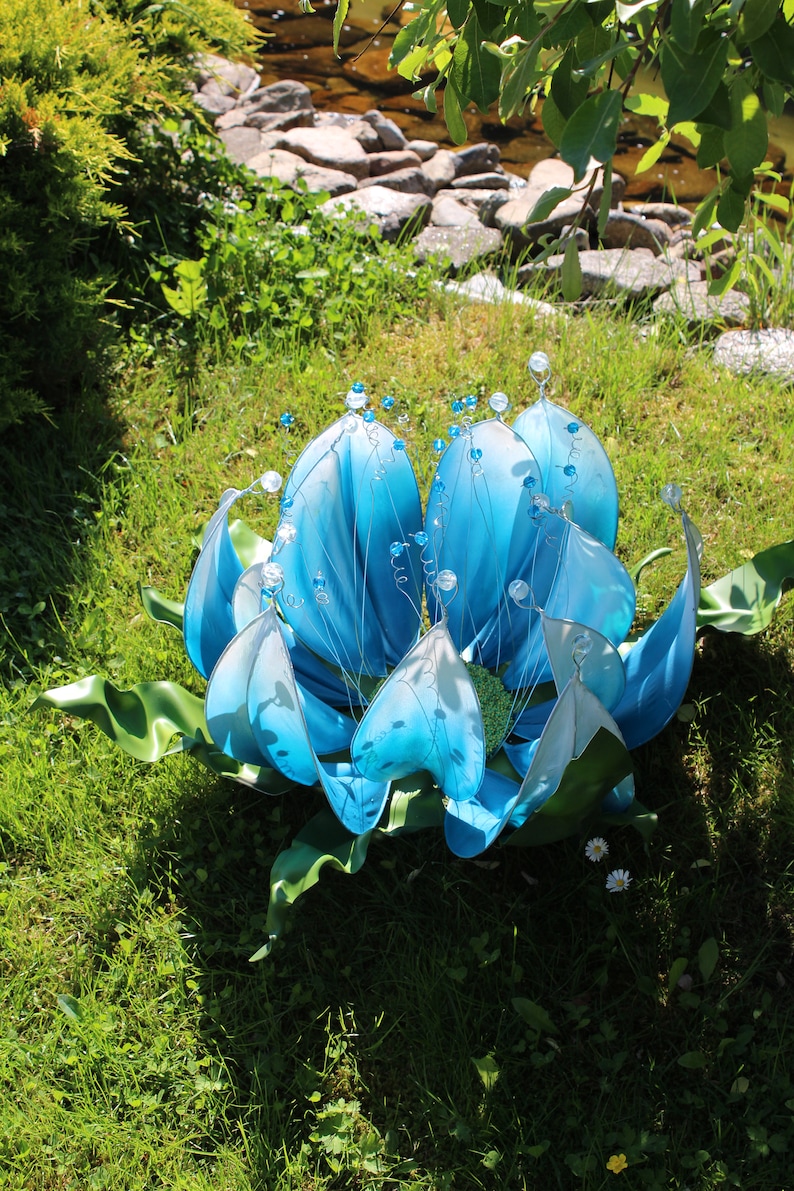 Pode incluir: Uma escultura de flor azul e branca com detalhes em contas. A escultura &eacute; feita de tecido e arame e est&aacute; sentada em um leito de grama verde.