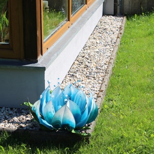 Pode incluir: Uma escultura de flor de metal azul com um centro branco e contas transparentes. A escultura est&aacute; sentada em um leito de cascalho ao lado de um edif&iacute;cio.