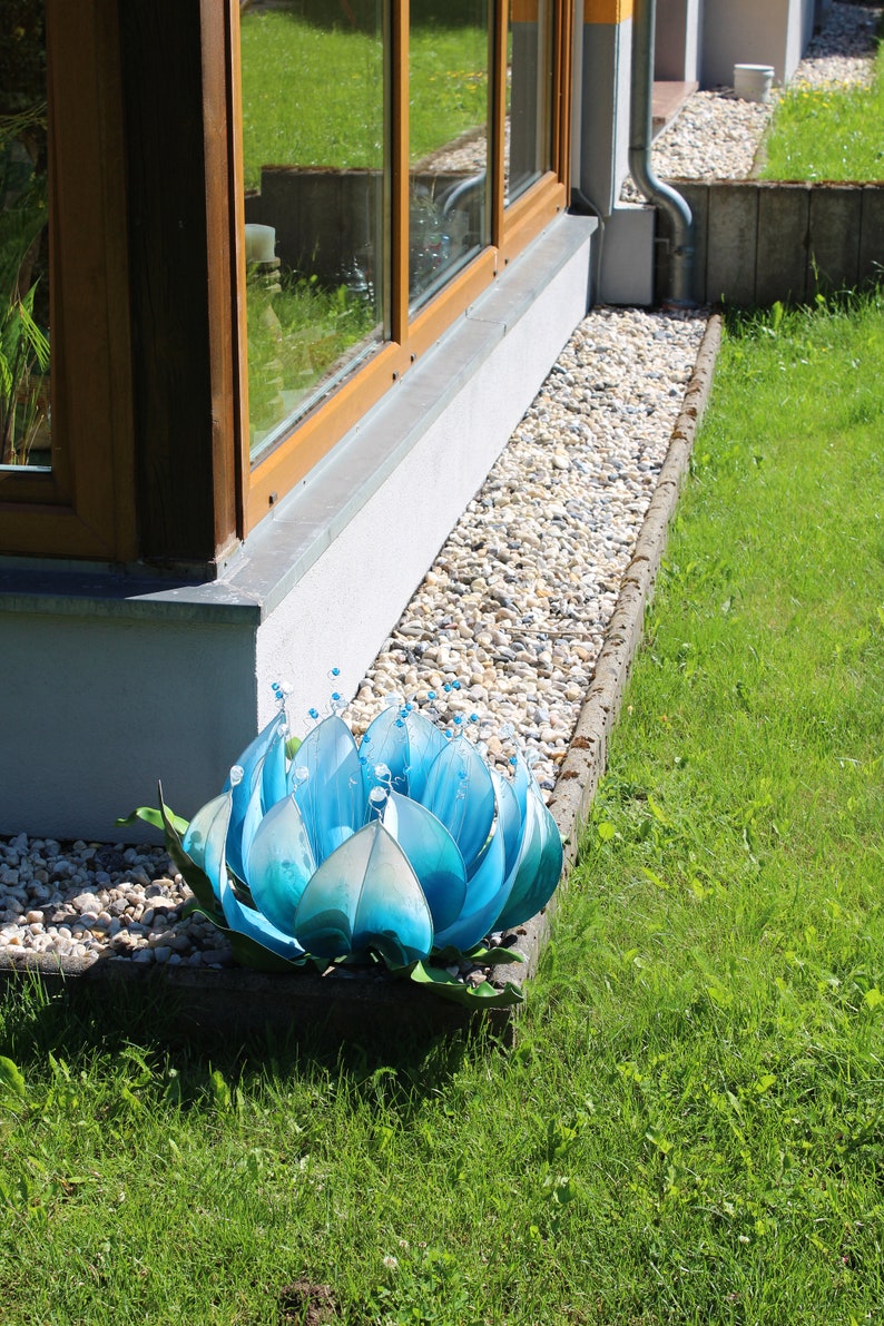 Pode incluir: Uma escultura de flor de metal azul com um centro branco e contas transparentes. A escultura est&aacute; sentada em um leito de cascalho ao lado de um edif&iacute;cio.