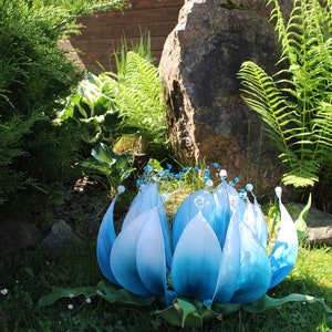 Pode incluir: Uma grande escultura de flor azul com detalhes brancos e contas de cristal. A escultura &eacute; feita de tecido e est&aacute; sentada em um leito de grama verde.