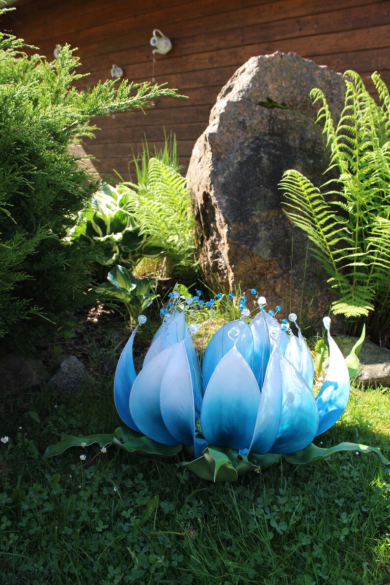 Pode incluir: Uma grande escultura de flor azul com detalhes brancos e contas de cristal. A escultura &eacute; feita de tecido e est&aacute; sentada em um leito de grama verde.