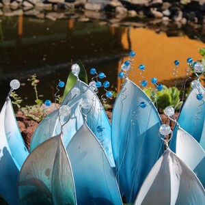 Pode incluir: Uma escultura de flor de vidro azul e branco com caules de contas. A escultura &eacute; feita de m&uacute;ltiplas camadas de p&eacute;talas de vidro e lembra um l&iacute;rio d'&aacute;gua.