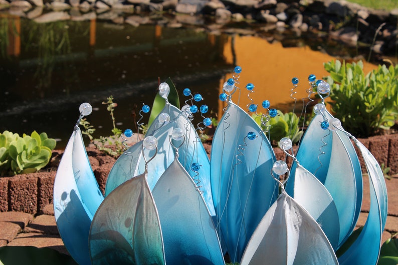 Pode incluir: Uma escultura de flor de vidro azul e branco com caules de contas. A escultura &eacute; feita de m&uacute;ltiplas camadas de p&eacute;talas de vidro e lembra um l&iacute;rio d'&aacute;gua.