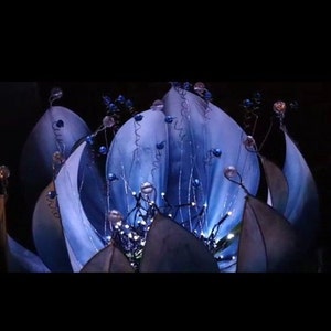 Pode incluir: Uma escultura de flor azul com luzes brancas e contas azuis. A flor &eacute; feita de tecido e tem um visual delicado e et&eacute;reo.