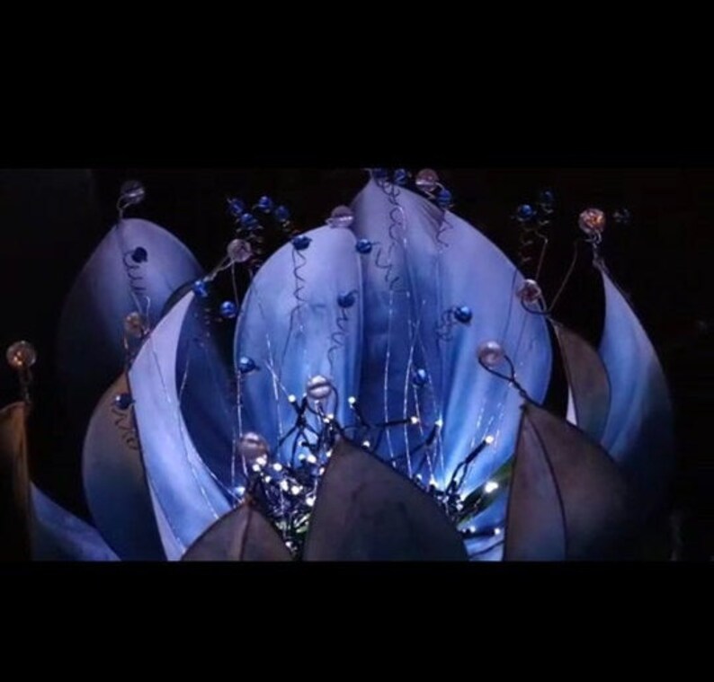 Pode incluir: Uma escultura de flor azul com luzes brancas e contas azuis. A flor &eacute; feita de tecido e tem um visual delicado e et&eacute;reo.