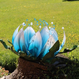 Pode incluir: Uma escultura de flor de vidro azul com detalhes brancos e contas de cristal, sentada em um toco de &aacute;rvore em um jardim gramado.
