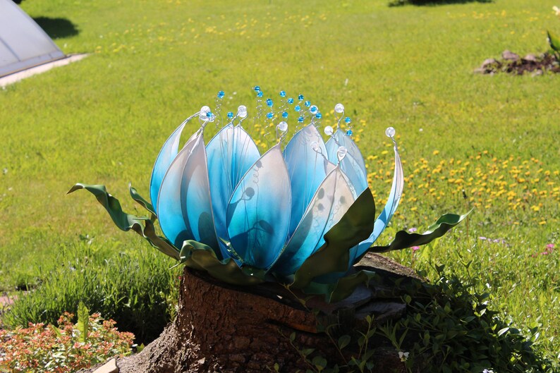 Pode incluir: Uma escultura de flor de vidro azul com detalhes brancos e contas de cristal, sentada em um toco de &aacute;rvore em um jardim gramado.