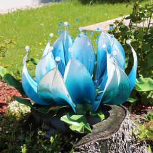 Pode incluir: Uma escultura de flor de metal azul e branco com um degradado de cor. A escultura est&aacute; colocada em um toco de &aacute;rvore em um jardim.
