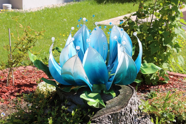 Pode incluir: Uma escultura de flor de metal azul e branco com um degradado de cor. A escultura est&aacute; colocada em um toco de &aacute;rvore em um jardim.