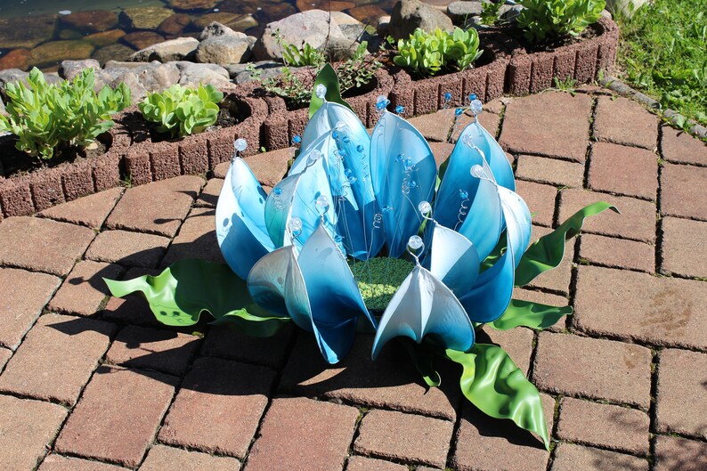 Pode incluir: Uma escultura de flor de metal azul e branco com folhas verdes. A escultura &eacute; feita de muitas pe&ccedil;as individuais de metal que s&atilde;o unidas para formar a flor.