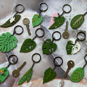 May include: A collection of 12 keychains featuring different leaf designs. The keychains are made of wood and painted in various shades of green, pink, and white. Each keychain has a metal ring and a small key attached.