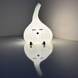 May include: A quirky white garlic-shaped lamp with a curved stem and a textured surface. The lamp has a cartoon face with closed eyes and small black feet. The lamp is illuminated, creating a soft glow and a reflection.