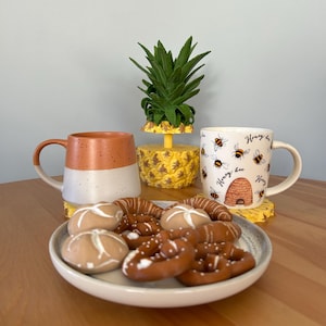 May include: Two mugs, one with a honey bee design and the other with a speckled brown and white glaze, sit on a wooden table with a plate of pretzels and cookies. A pineapple-shaped ceramic planter with a green plant sits between the mugs.