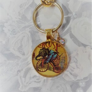 May include: Gold-tone keychain featuring a round charm with a vintage illustration of an elephant riding a tricycle. The charm has the word "Circus" printed on it.