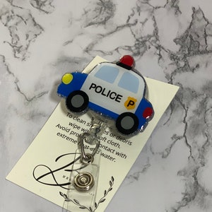 May include: A blue and white police car badge reel with a silver clip and a clear plastic holder. The badge reel has the word "POLICE" and the letter "P" on it.