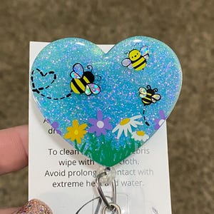 Puede incluir: Un portagafete en forma de coraz&oacute;n azul con purpurina, con tres abejas de dibujos animados y flores. El portagafete tiene un carrete retr&aacute;ctil plateado.