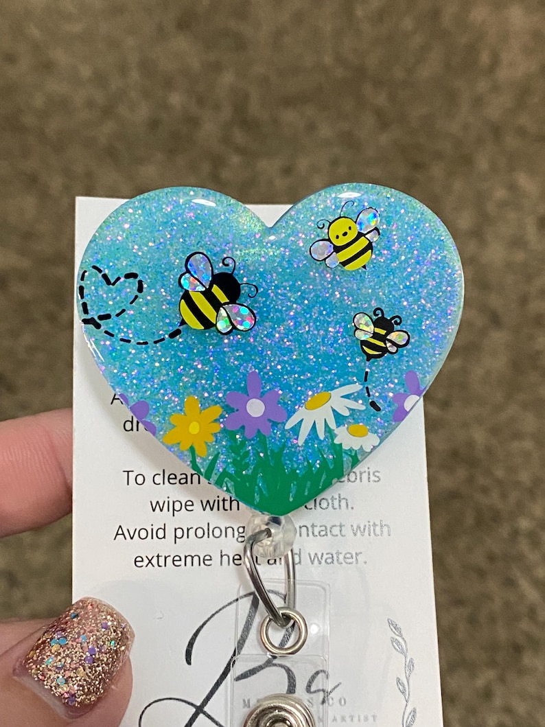 Puede incluir: Un portagafete en forma de coraz&oacute;n azul con purpurina, con tres abejas de dibujos animados y flores. El portagafete tiene un carrete retr&aacute;ctil plateado.