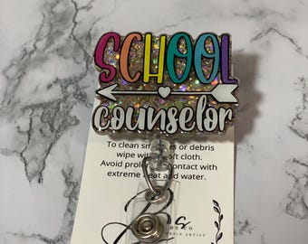 May include: A glittery badge reel with the words "SCHOOL COUNSELOR" in colorful letters. The badge has a white arrow and heart design. The reel is attached to a clear plastic holder.