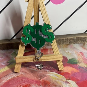 May include: A green and silver dollar sign badge reel. The badge reel features three dollar signs with a glittery silver center and a green outline. The badge reel is displayed on a small wooden easel.