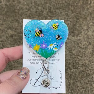 Puede incluir: Un portagafete en forma de coraz&oacute;n azul con purpurina con un dise&ntilde;o de abejas y flores. El texto en la parte posterior dice: "Para limpiar, limpiar con un pa&ntilde;o. Evite el contacto prolongado con calor extremo y agua."