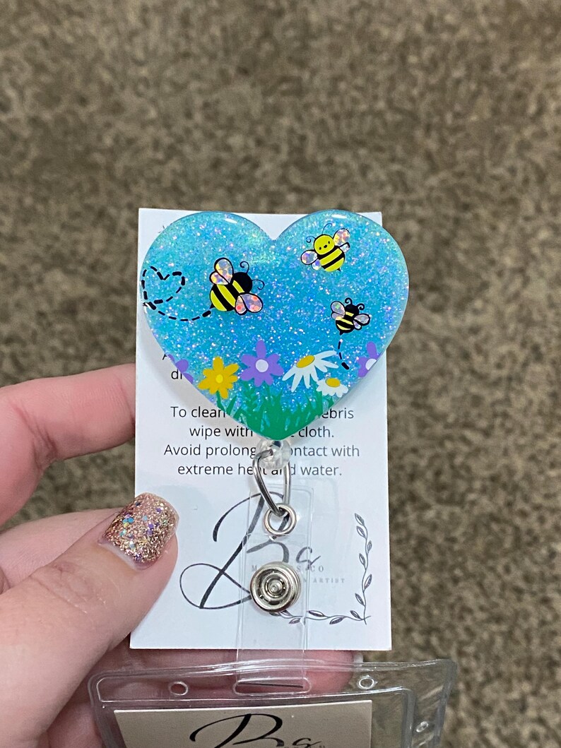 Puede incluir: Un portagafete en forma de coraz&oacute;n azul con purpurina con un dise&ntilde;o de abejas y flores. El texto en la parte posterior dice: "Para limpiar, limpiar con un pa&ntilde;o. Evite el contacto prolongado con calor extremo y agua."
