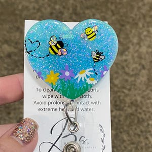 Puede incluir: Un portagafete en forma de coraz&oacute;n azul con purpurina con un dise&ntilde;o de abejas y flores. El portagafete tiene un respaldo de pl&aacute;stico transparente y un clip de metal. El texto en la tarjeta dice "Para limpiar, limpiar con un pa&ntilde;o. Evite el contacto prolongado con calor extremo y agua."