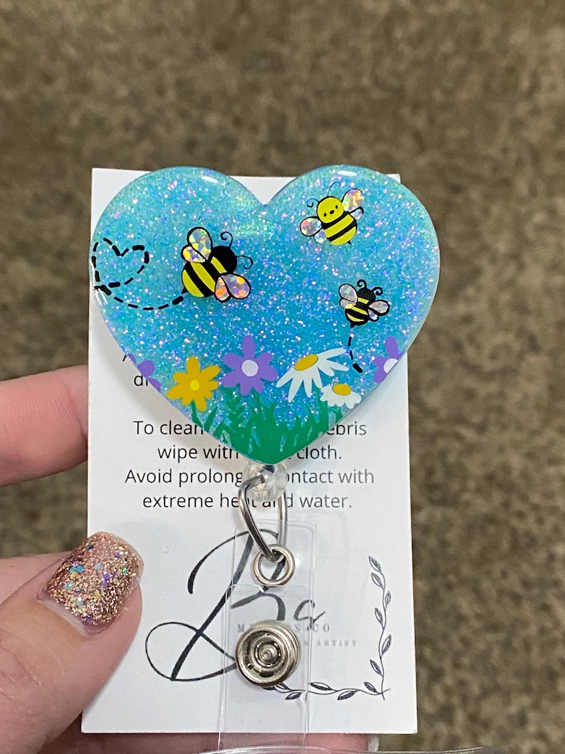Puede incluir: Un portagafete en forma de coraz&oacute;n azul con purpurina con un dise&ntilde;o de abejas y flores. El portagafete tiene un respaldo de pl&aacute;stico transparente y un clip de metal. El texto en la tarjeta dice "Para limpiar, limpiar con un pa&ntilde;o. Evite el contacto prolongado con calor extremo y agua."