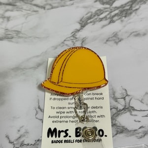 May include: A yellow hard hat badge reel with a glittery gold border. The badge reel has the text "Mrs. Boo. BADGE REELS FOR EVERYDAY!" on a white card. The badge reel is attached to a clear plastic holder.