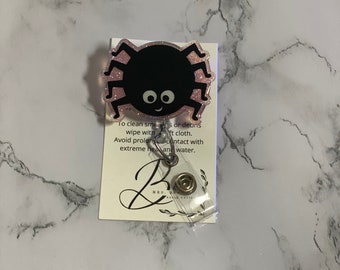 May include: A black spider-shaped badge reel with a pink glitter border and a smiling face. The badge reel has a clear plastic retractable cord and a metal clip. The text on the attached card reads "To clean smudges or debris wipe with a soft cloth."