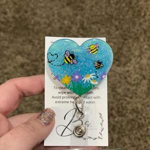 Puede incluir: Un soporte para credenciales de resina azul en forma de coraz&oacute;n con purpurina y un dise&ntilde;o de abejas y flores. El texto en la parte posterior dice "Para limpiar, limpiar con un pa&ntilde;o h&uacute;medo. Evite el contacto prolongado con el calor extremo y el agua."