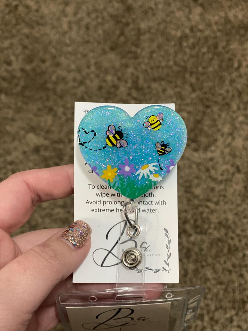 Puede incluir: Un soporte para credenciales de resina azul en forma de coraz&oacute;n con purpurina y un dise&ntilde;o de abejas y flores. El texto en la parte posterior dice "Para limpiar, limpiar con un pa&ntilde;o h&uacute;medo. Evite el contacto prolongado con el calor extremo y el agua."