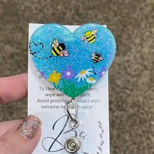 Puede incluir: Un portagafete azul en forma de coraz&oacute;n con brillantina y un dise&ntilde;o de abeja. El portagafete tiene una correa de pl&aacute;stico transparente y un clip de metal. El texto en la tarjeta dice: "Para limpiar, limpiar con un pa&ntilde;o h&uacute;medo. Evite el contacto prolongado con el calor extremo y el agua."