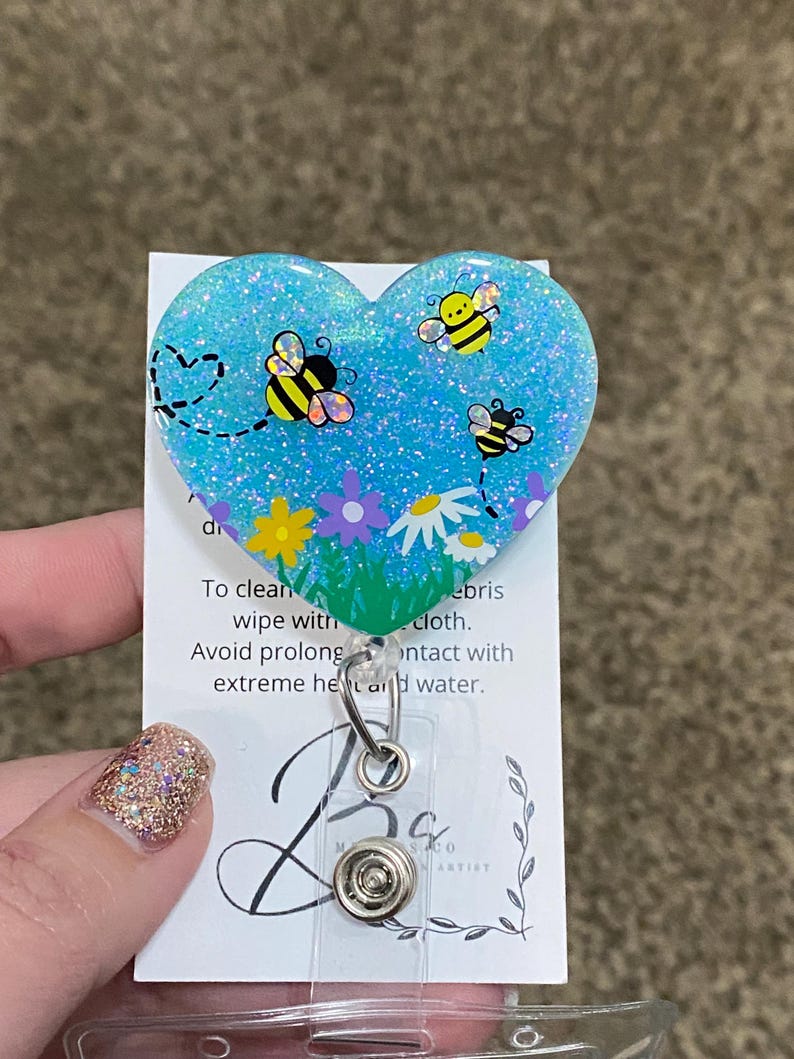 Puede incluir: Un portagafete azul en forma de coraz&oacute;n con brillantina y un dise&ntilde;o de abeja. El portagafete tiene una correa de pl&aacute;stico transparente y un clip de metal. El texto en la tarjeta dice: "Para limpiar, limpiar con un pa&ntilde;o h&uacute;medo. Evite el contacto prolongado con el calor extremo y el agua."