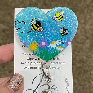 Puede incluir: Un portagafete en forma de coraz&oacute;n azul con purpurina con un dise&ntilde;o de abejas y flores. El portagafete tiene un clip de pl&aacute;stico transparente y un pivote de metal.