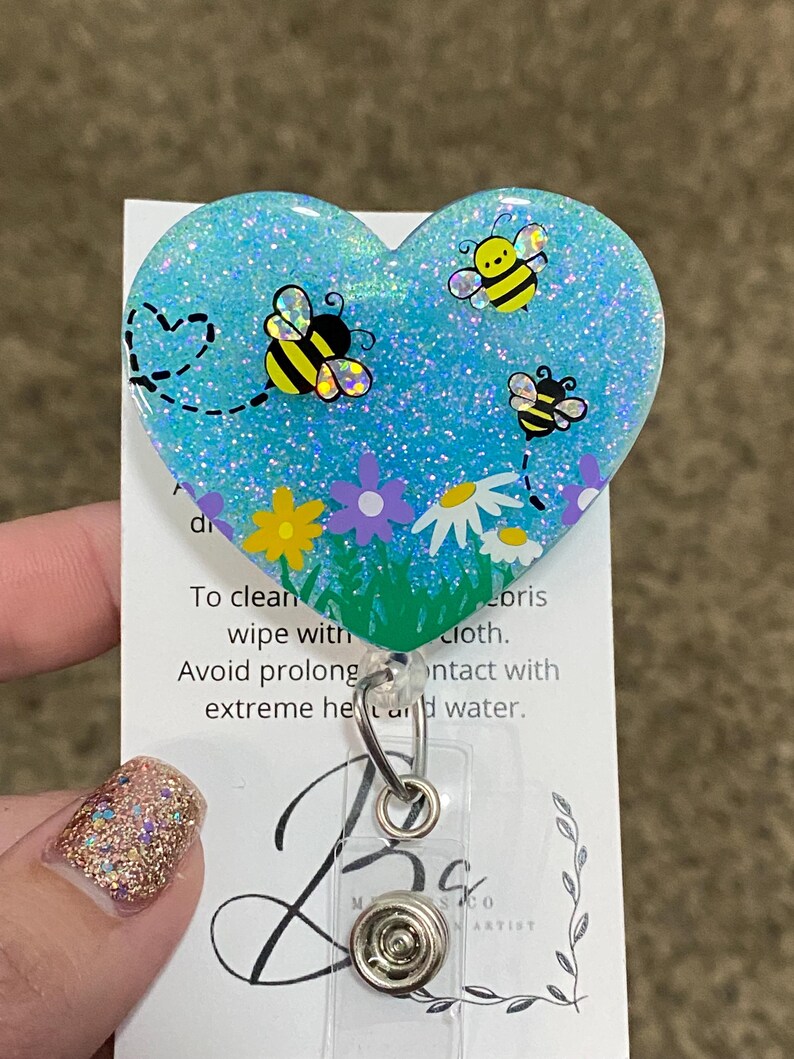 Puede incluir: Un portagafete en forma de coraz&oacute;n azul con purpurina con un dise&ntilde;o de abejas y flores. El portagafete tiene un clip de pl&aacute;stico transparente y un pivote de metal.