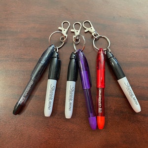 May include: Five keychains with different colored markers attached. The markers are black, white, purple, red, and black. The markers are labeled "Sharpie Mini" and "Mr. Sketch".