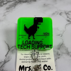May include: A green badge reel with a black pixelated dinosaur graphic and the text "LOADING... TECH SUPPORT" and "404". The badge reel also has a clear plastic strap and a metal clip. The text "Mrs. Co. BADGE REELS FOR EVERYDAY!" is also visible.