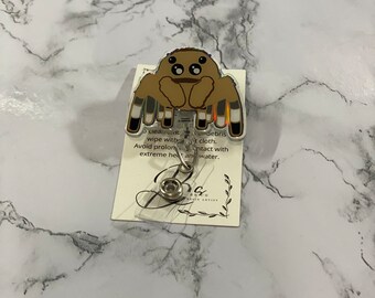 May include: A brown spider-shaped badge reel with a cartoon design. The badge reel has a clear plastic covering and a retractable cord. The attached card has text that reads: "To clean...wipe with...Avoid prolonged...extreme heat and water."