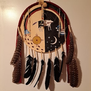 May include: A Native American-style dreamcatcher with a circular design. The dreamcatcher features a painted split design with a sun symbol, a wolf, and a crescent moon. It is adorned with black feathers and brown feathers, and red ribbon.