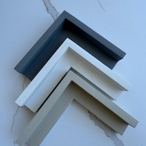 May include: Three L-shaped corner sections of picture frames stacked vertically. The top frame is dark charcoal gray, the middle frame is matte white, and the bottom frame is a light beige, all featuring a smooth, clean-lined profile design.
