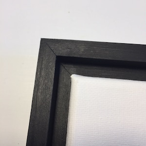 May include: Close-up of the corner of a black wood frame surrounding a white canvas.
