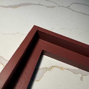 May include: A close-up of a burgundy-colored wooden picture frame corner. The frame has a simple, rectangular design with a matte finish. The background is a white surface with subtle, marbled patterns.