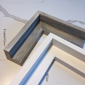May include: Two picture frames, one in white and one in a light gray color called "Coastal Grey". The frames are made of wood and have a simple, modern design.