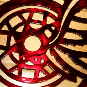 May include: A close-up of a red metal wall art piece with a circular design and intricate cut-out patterns. The artwork has a glossy finish and is mounted on a light-colored wooden surface. The design features a central circle and wing-like shapes.